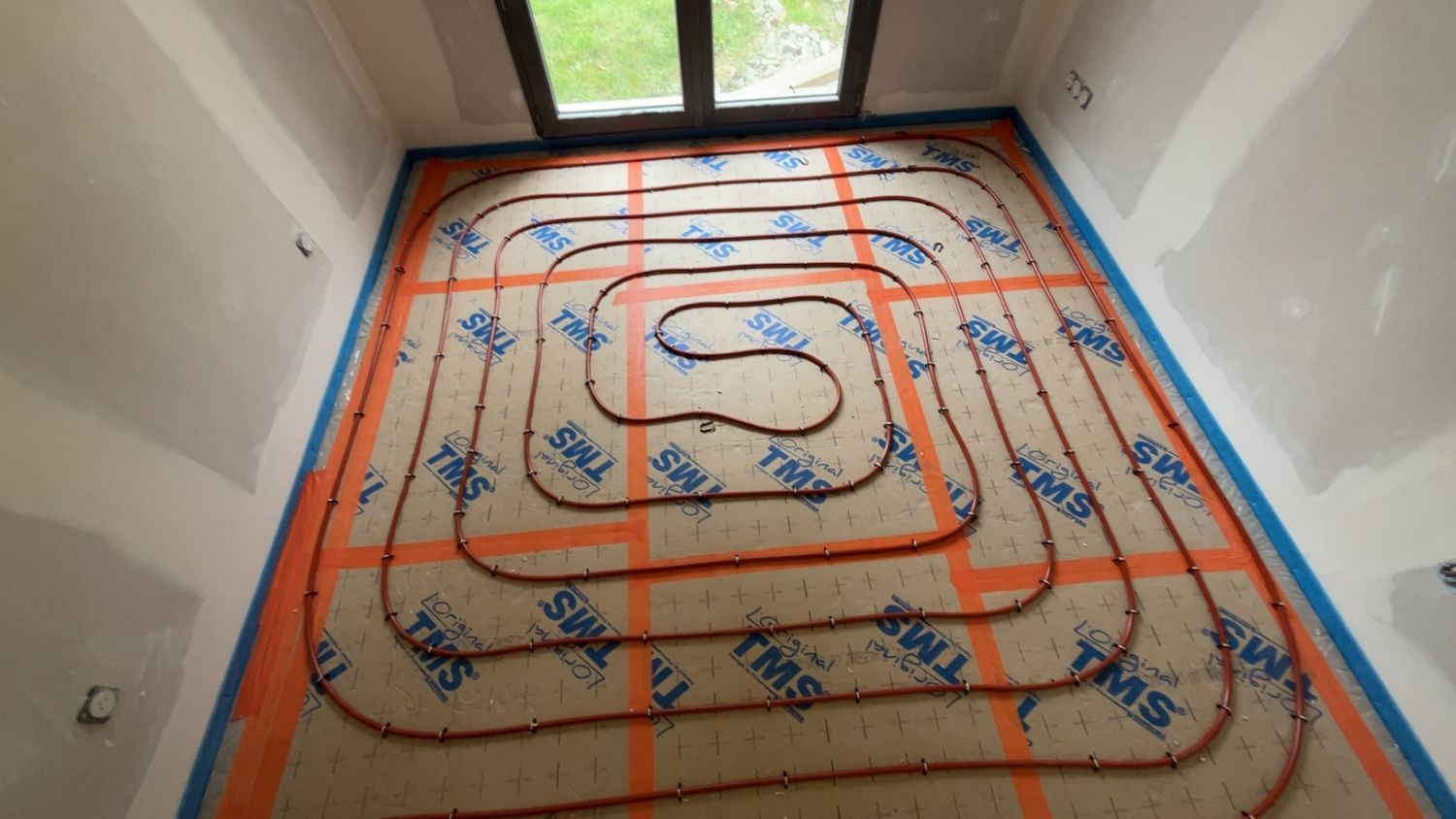 Installation d'un système de chauffage au sol avec des tuyaux orange posés sur une grille, dans une pièce avec fenêtre.