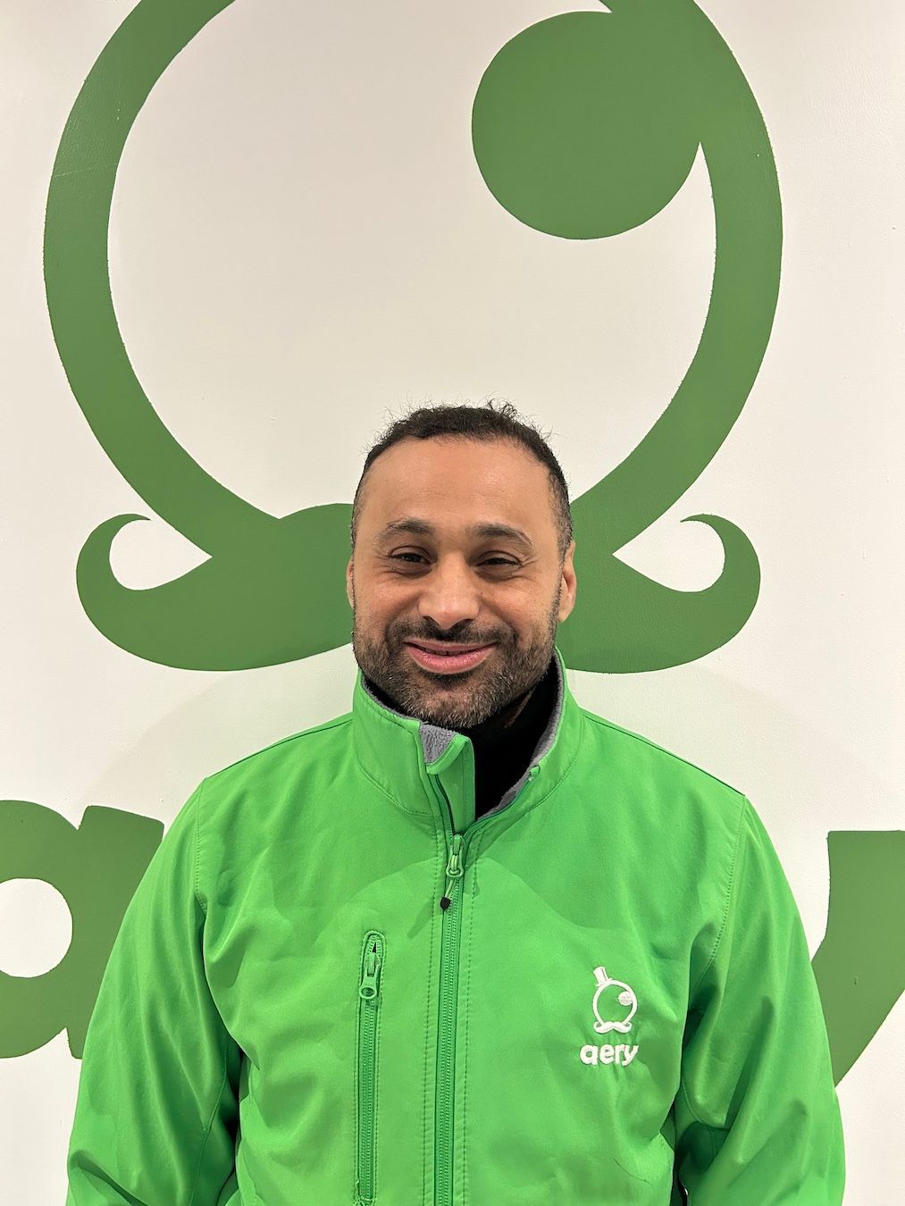Un homme en veste verte sourit devant un logo vert sur un mur blanc.