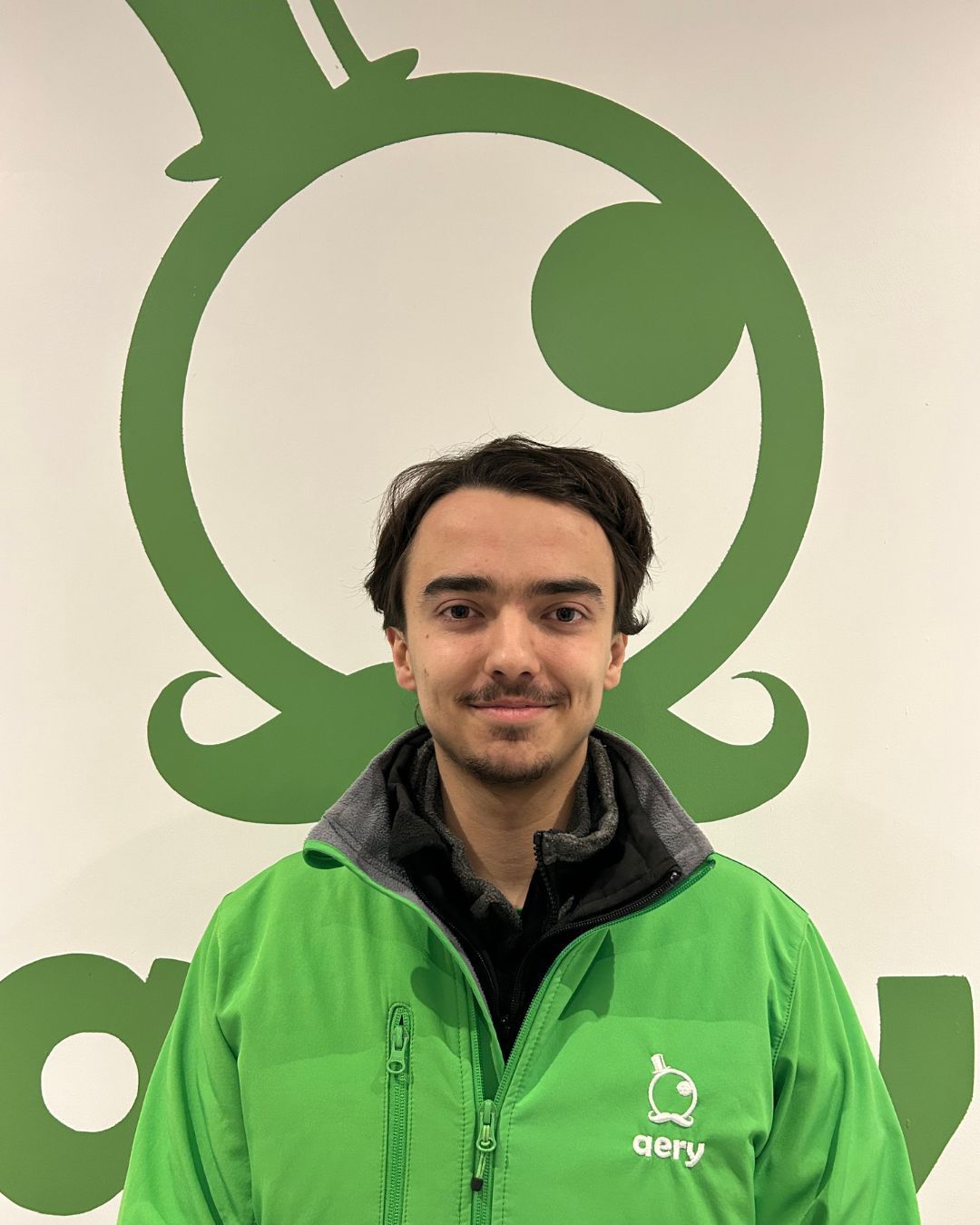 Un homme portant une veste verte sourit devant un logo d'entreprise vert.