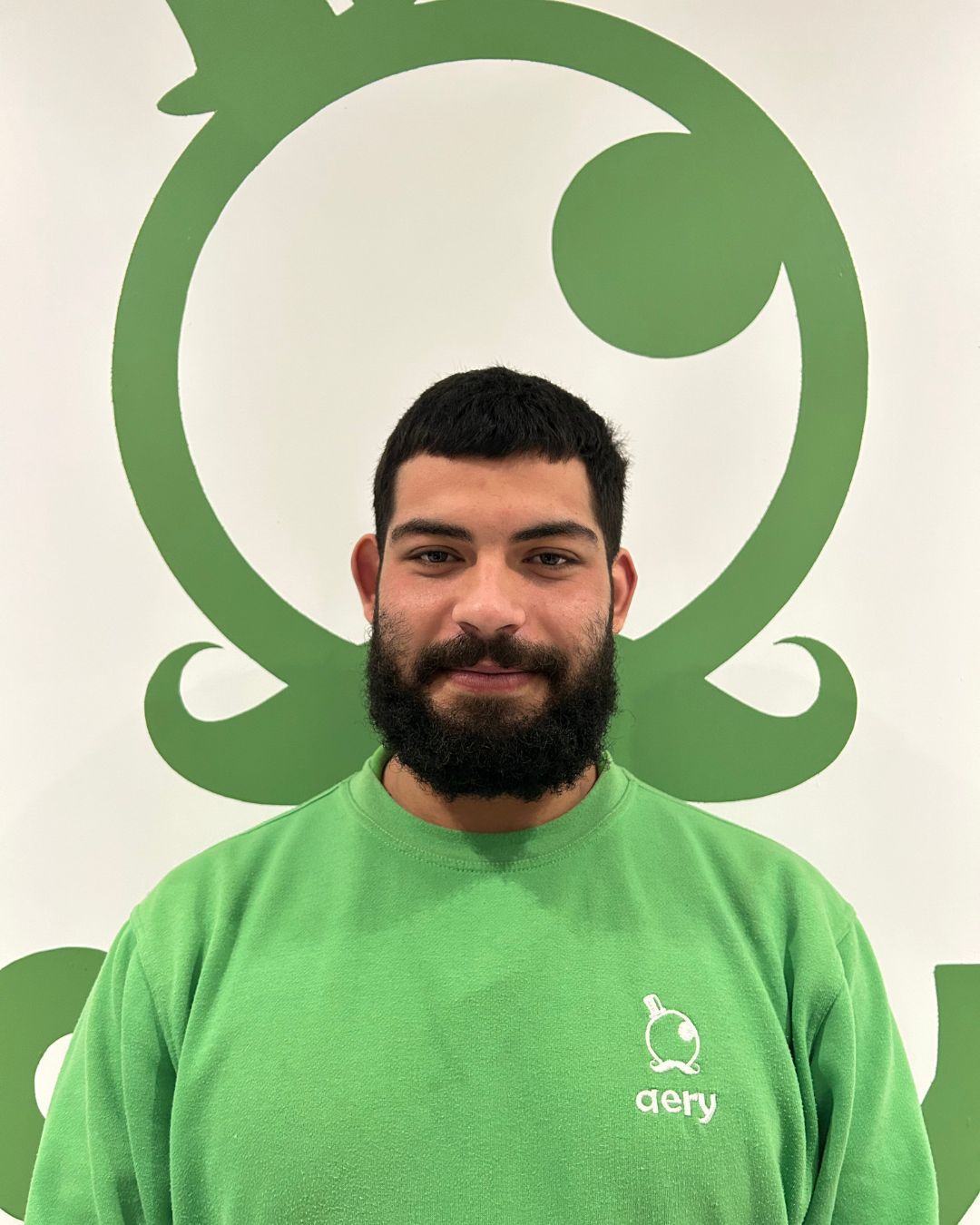 Un homme barbu portant une chemise verte se tient devant un logo vert sur un mur blanc.