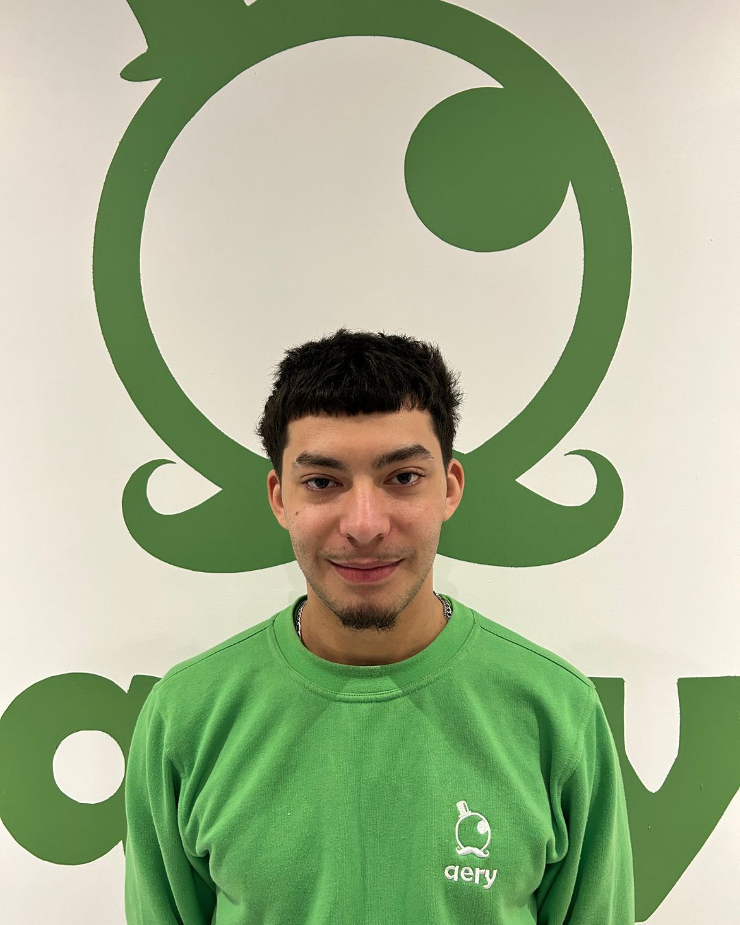 Une personne en pull vert sourit devant un logo vert sur un mur blanc.