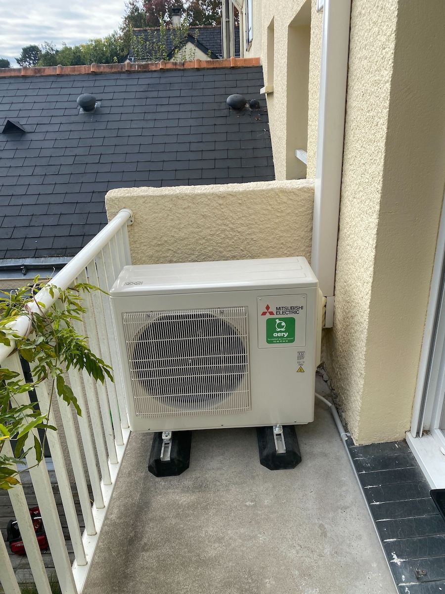 Unité de climatisation sur un balcon. L'unité blanche repose sur des supports noirs, contre un mur.