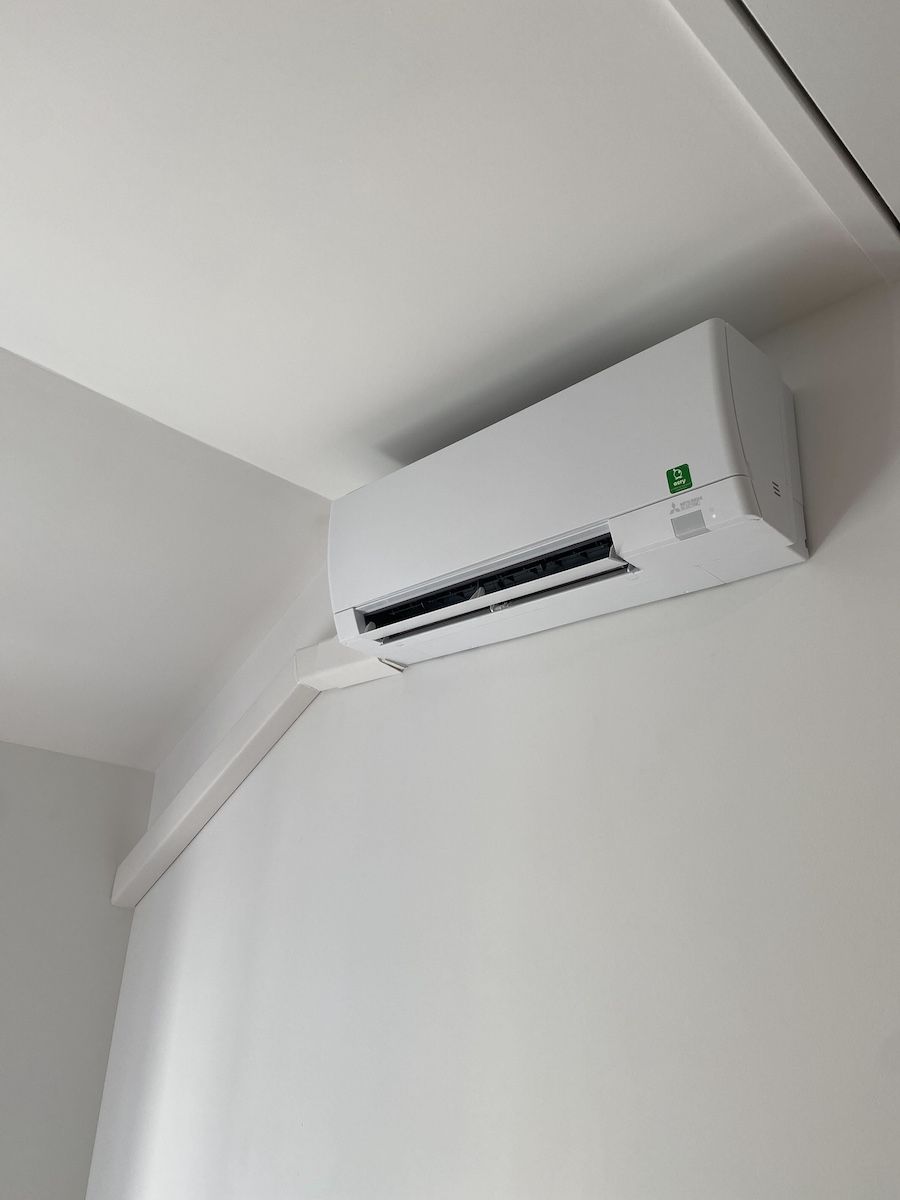 Climatiseur mural blanc. Grilles d'aération noires. Fixé au mur blanc près du plafond.