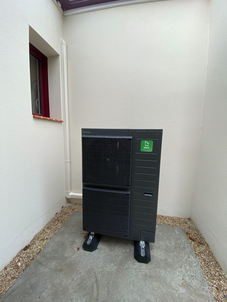 Unité de pompe à chaleur de couleur sombre installée à l'extérieur entre deux murs blancs, à côté d'une fenêtre.