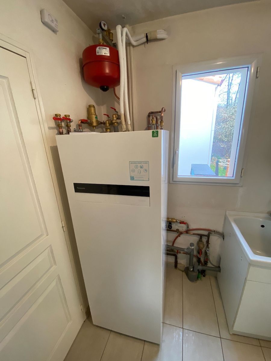 Pompe à chaleur blanche et vase d'expansion rouge dans une buanderie avec une petite fenêtre et un évier.