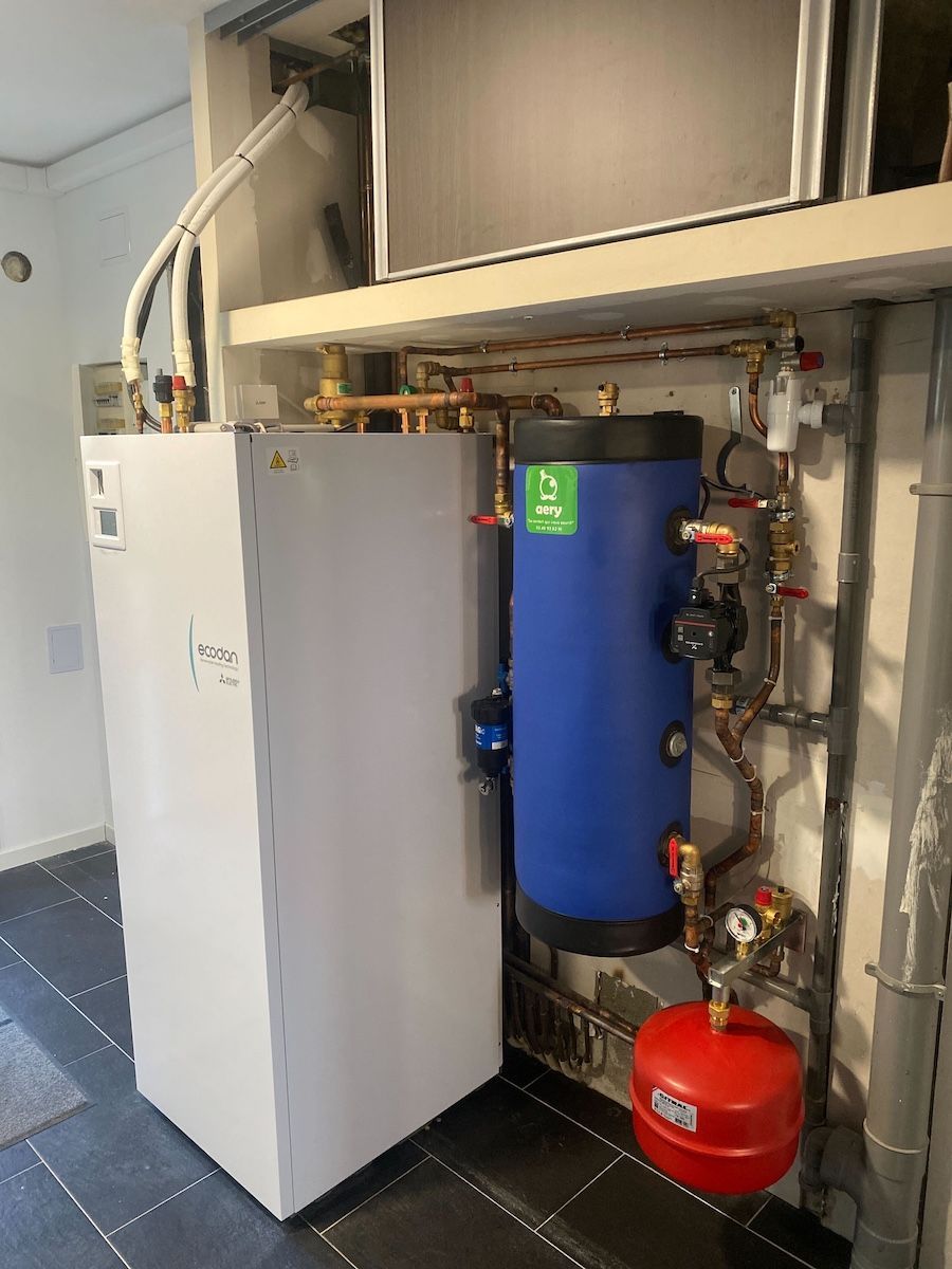 Un appareil de chauffage blanc se trouve à côté d'un réservoir d'eau muni d'un vase d'expansion rouge, dans une buanderie.