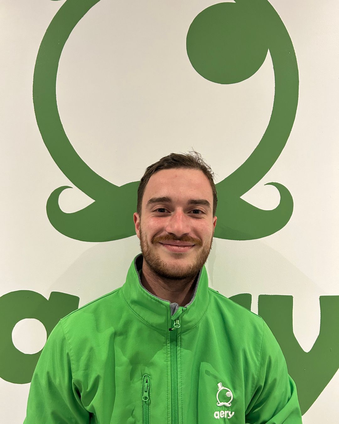 Un homme portant une veste verte sourit devant un logo vert sur un mur blanc.