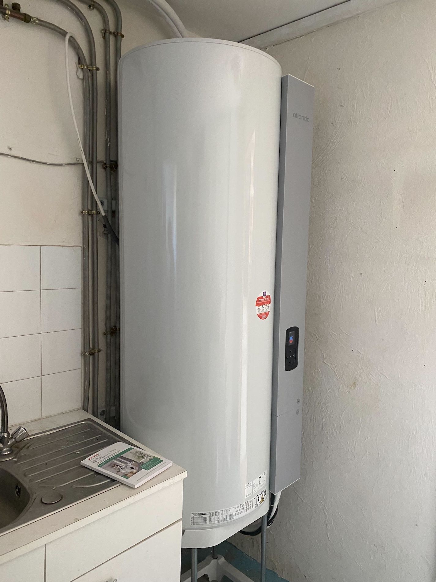 Chauffe-eau blanc à côté d'un évier, tuyaux au mur.