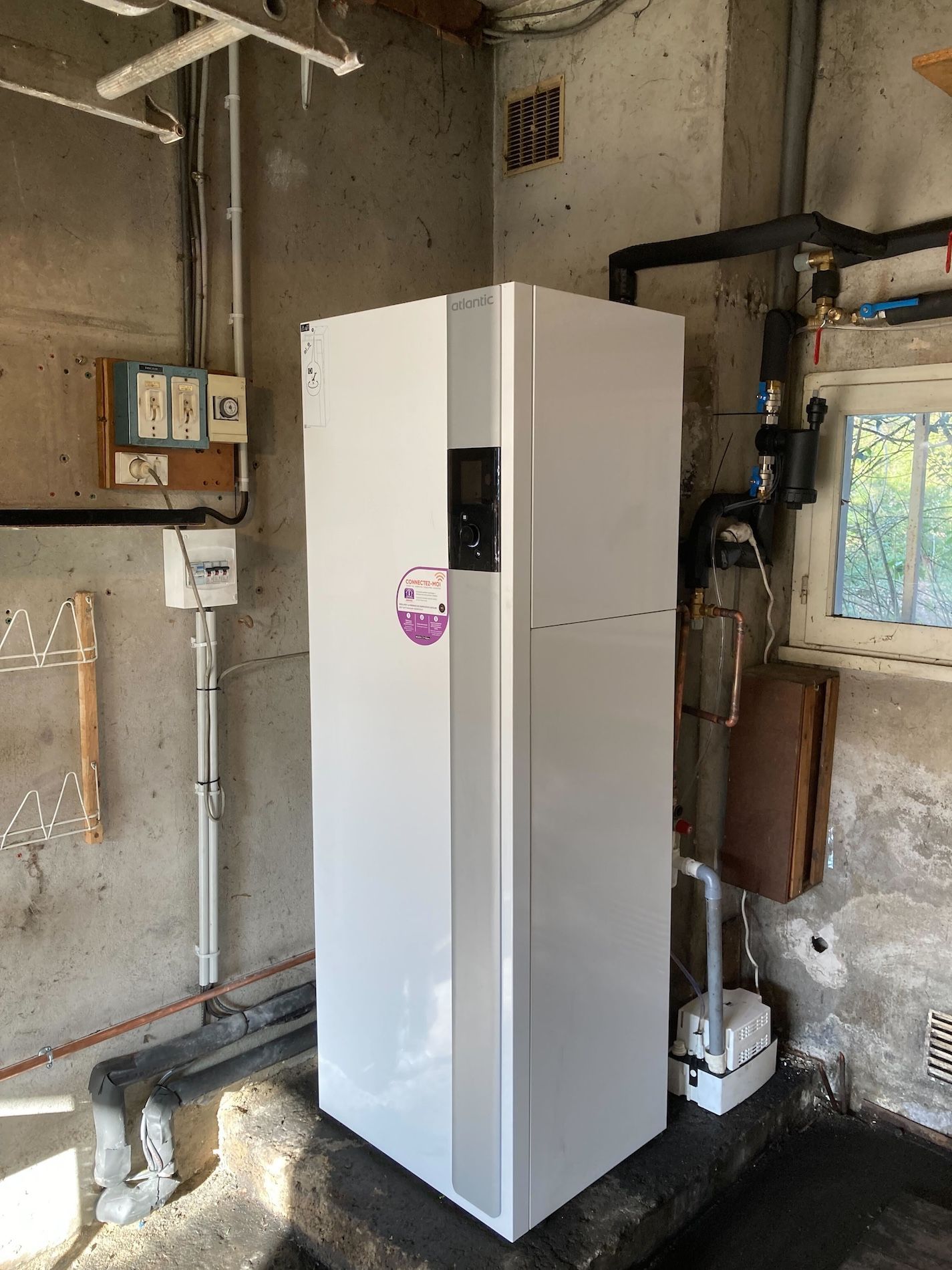 Unité de pompe à chaleur blanche dans une buanderie, avec tuyaux et petite fenêtre visibles.