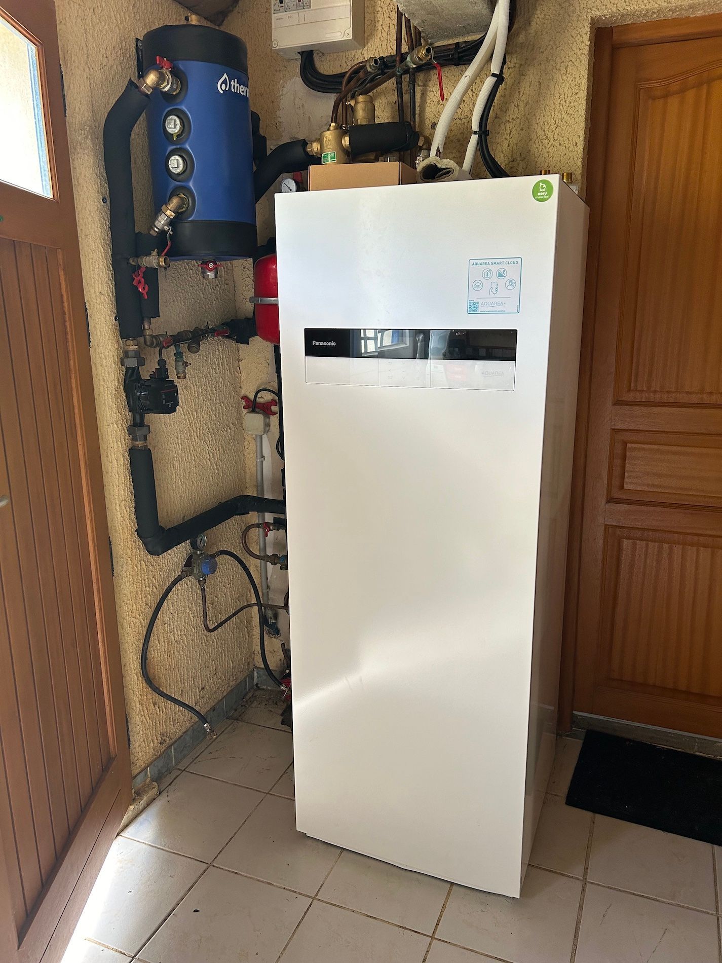 Unité de pompe à chaleur blanche dans une buanderie, à côté d'un réservoir bleu et d'une porte en bois.