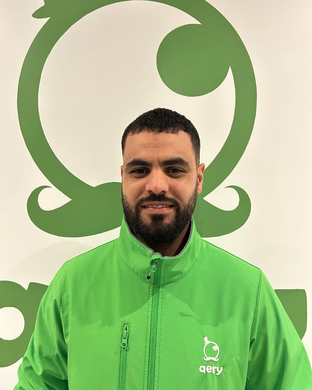 Un homme en veste verte sourit, debout devant un logo vert et blanc.