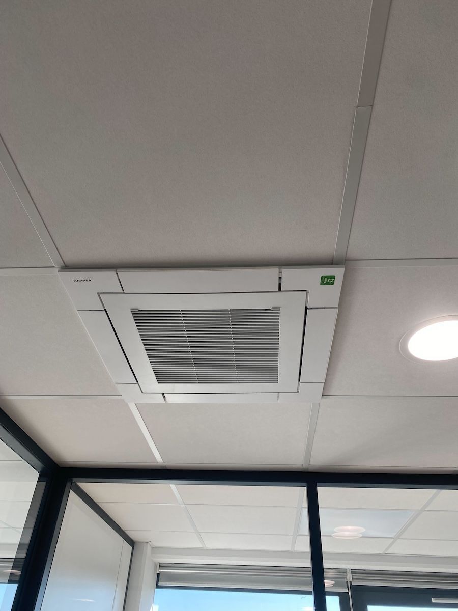 Climatiseur fixé au plafond dans un bâtiment commercial avec des dalles blanches et une vue sur des fenêtres en verre.