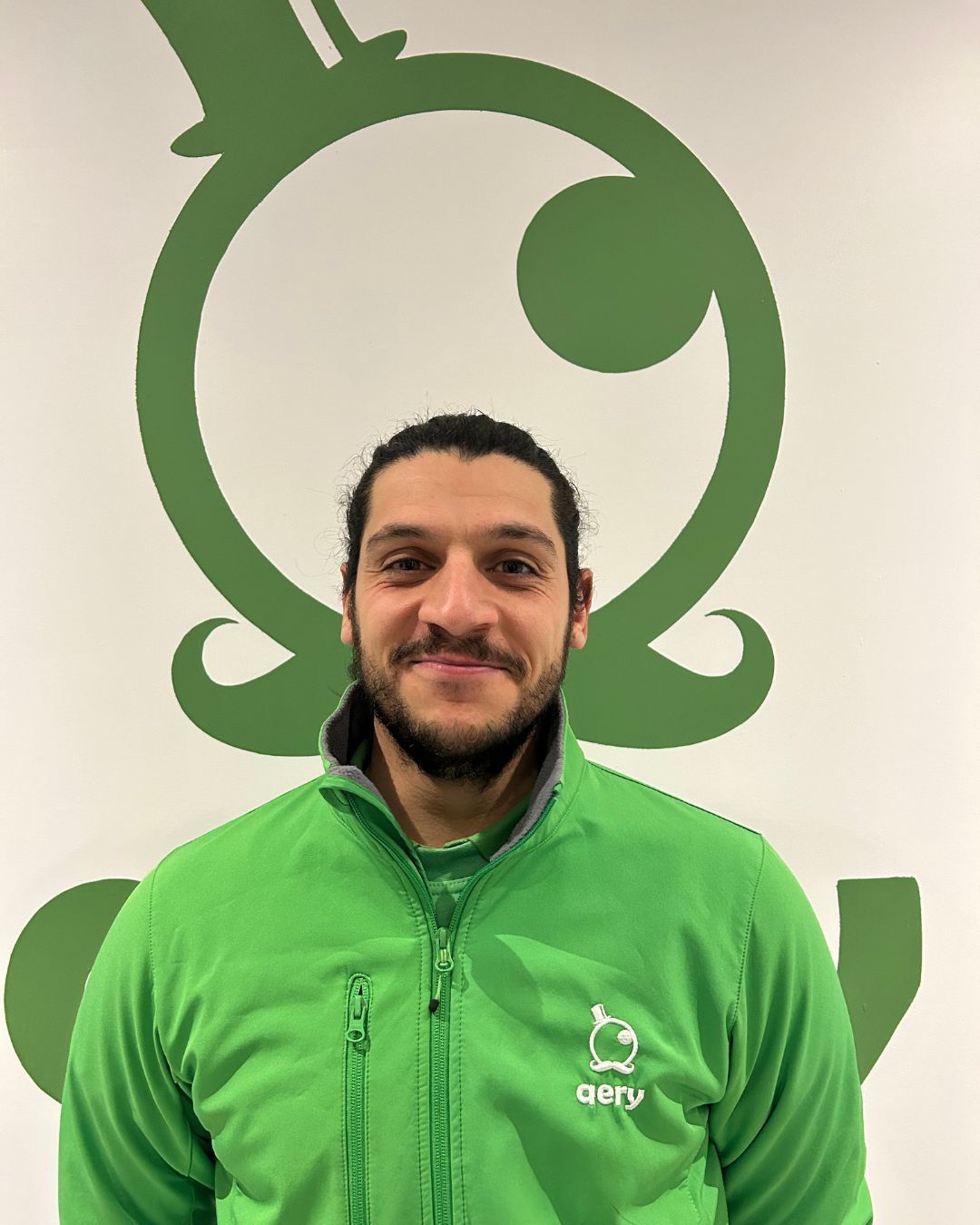 Un homme en veste verte sourit devant le logo vert d'une entreprise.