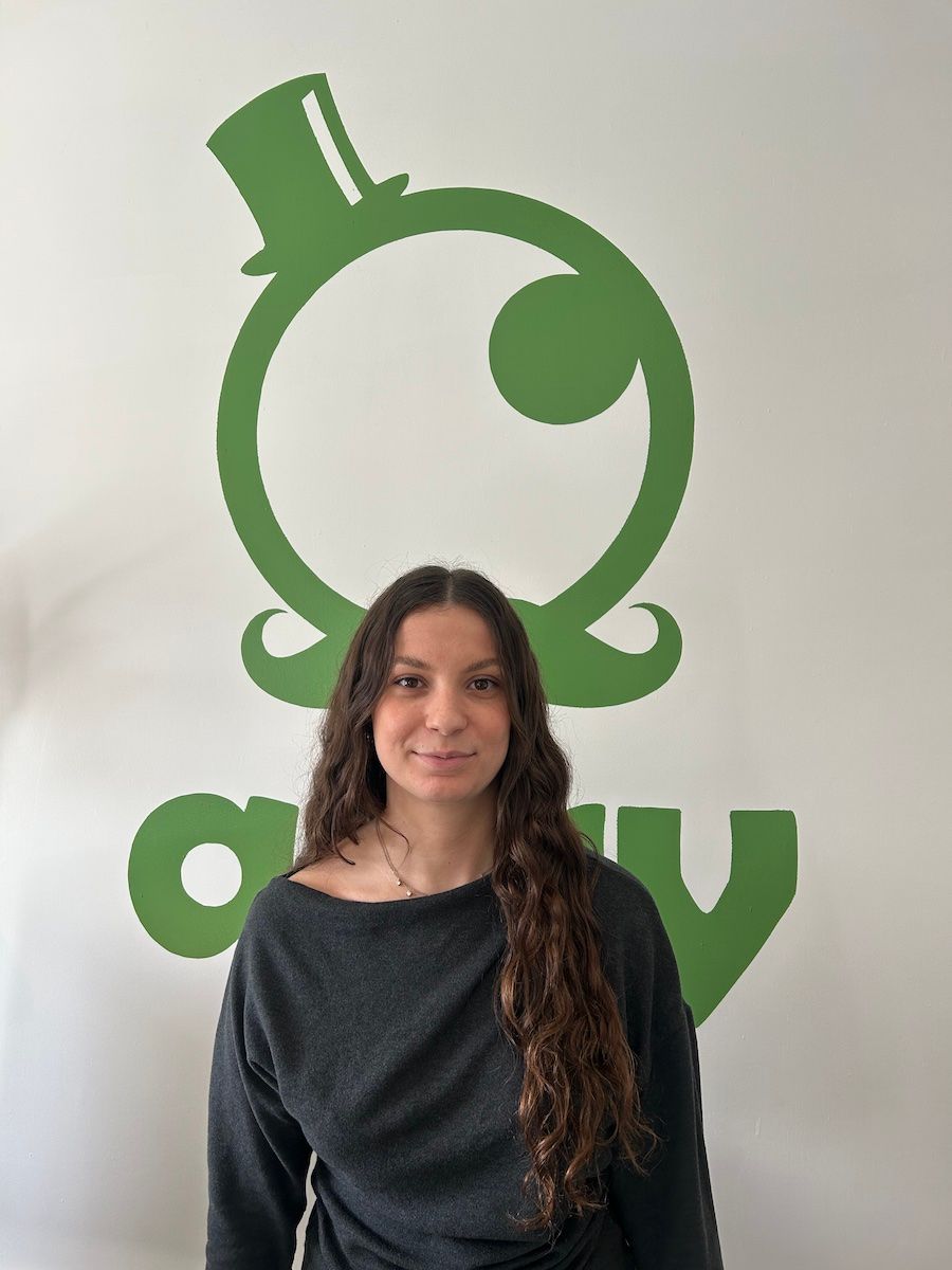 Femme aux longs cheveux bouclés, vêtue d'une chemise noire à manches longues, debout devant un mur bleu.