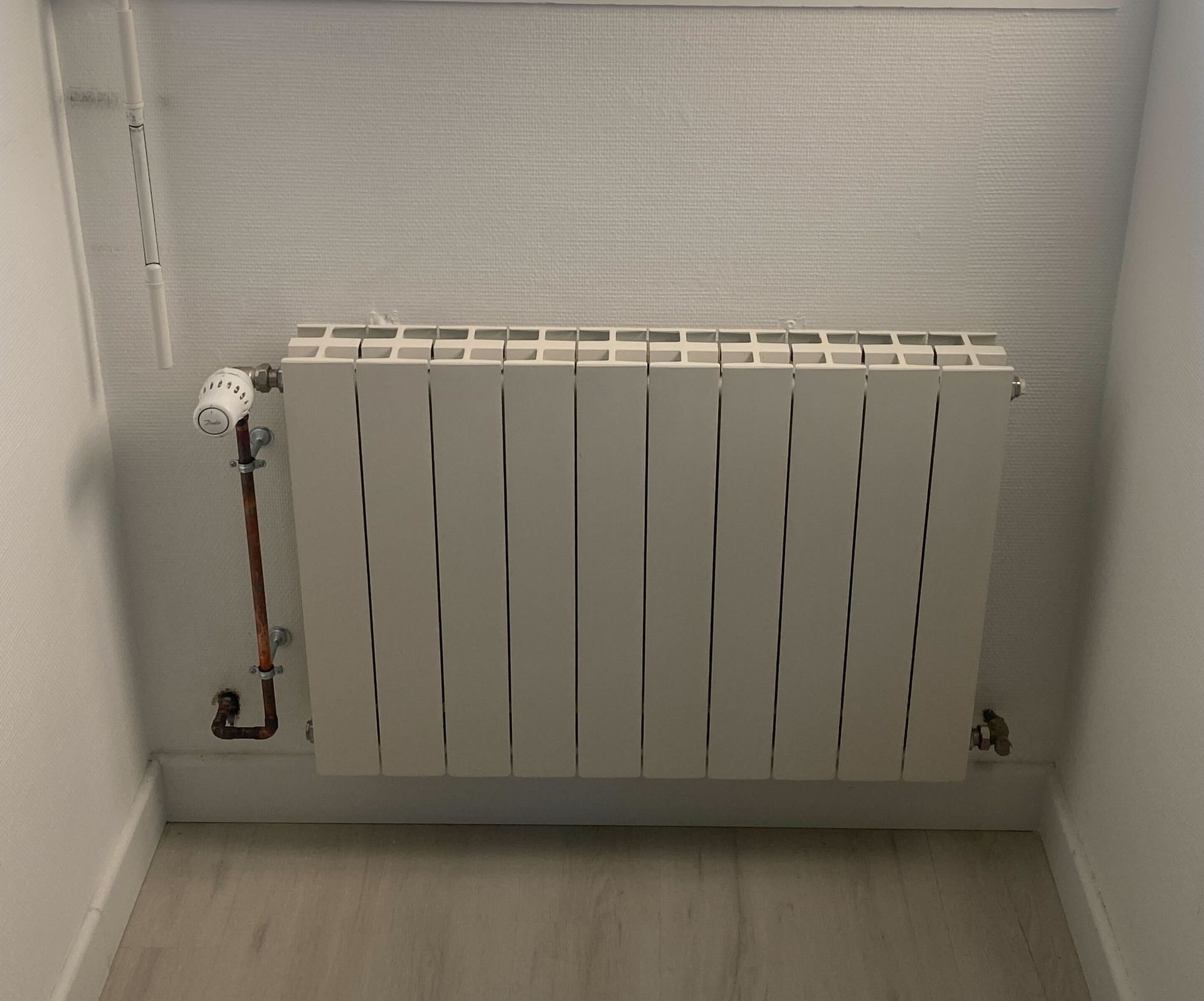 Radiateur blanc fixé au mur d'une pièce, avec des tuyaux en cuivre et un thermostat.