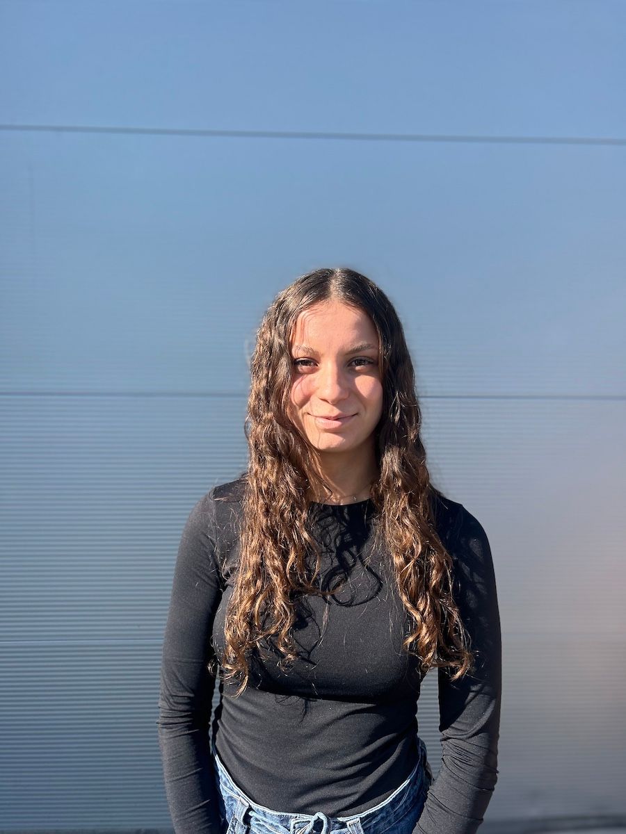 Femme aux longs cheveux bouclés, vêtue d'une chemise noire à manches longues, debout devant un mur bleu.