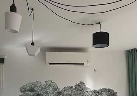 Climatiseur fixé sur un mur blanc, avec des lampes suspendues et une fresque représentant un paysage forestier.