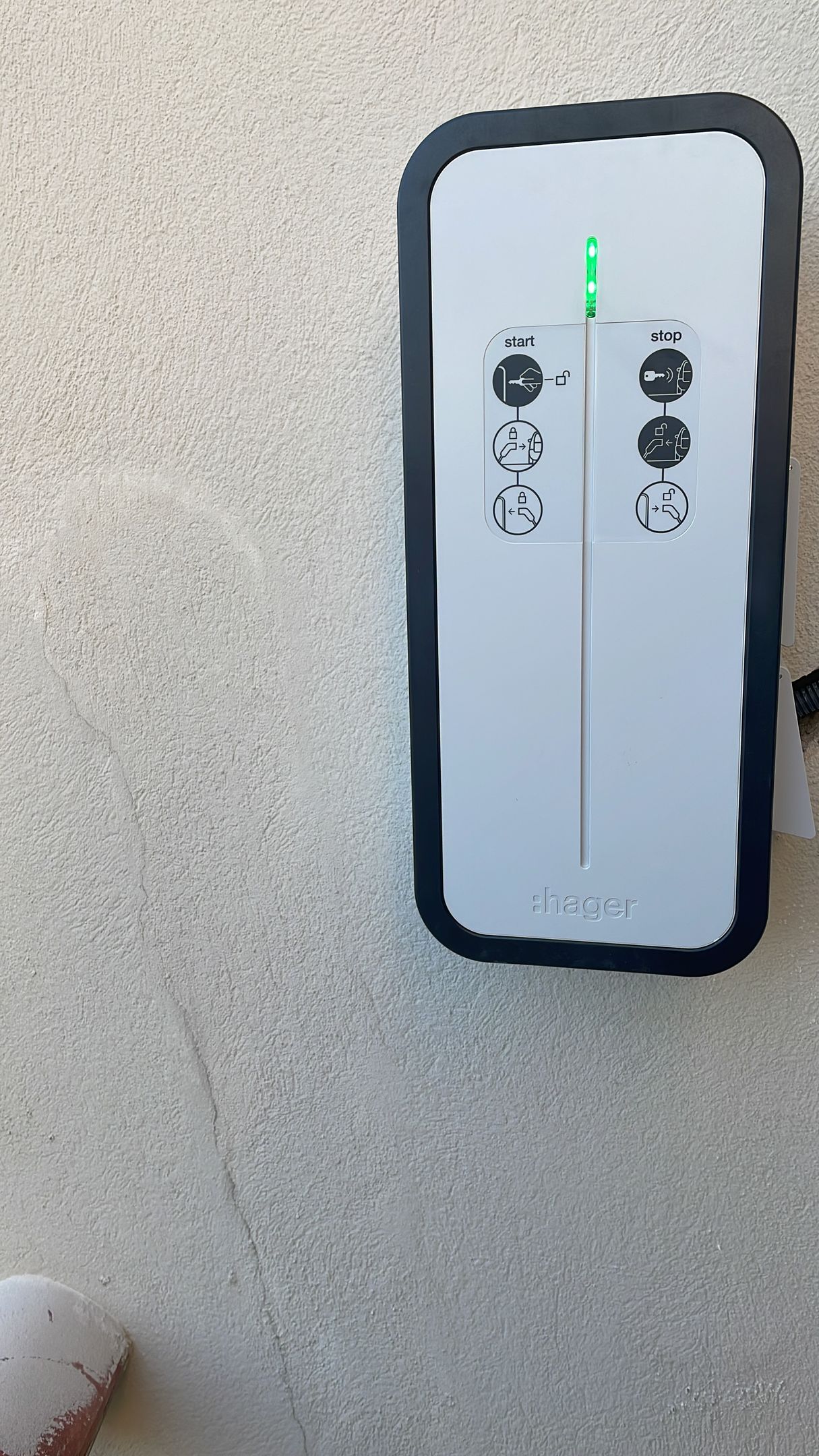 Borne de recharge murale pour véhicule électrique avec câble enroulé, fixée sur un mur de briques à côté d'une porte de garage et d'une voiture.