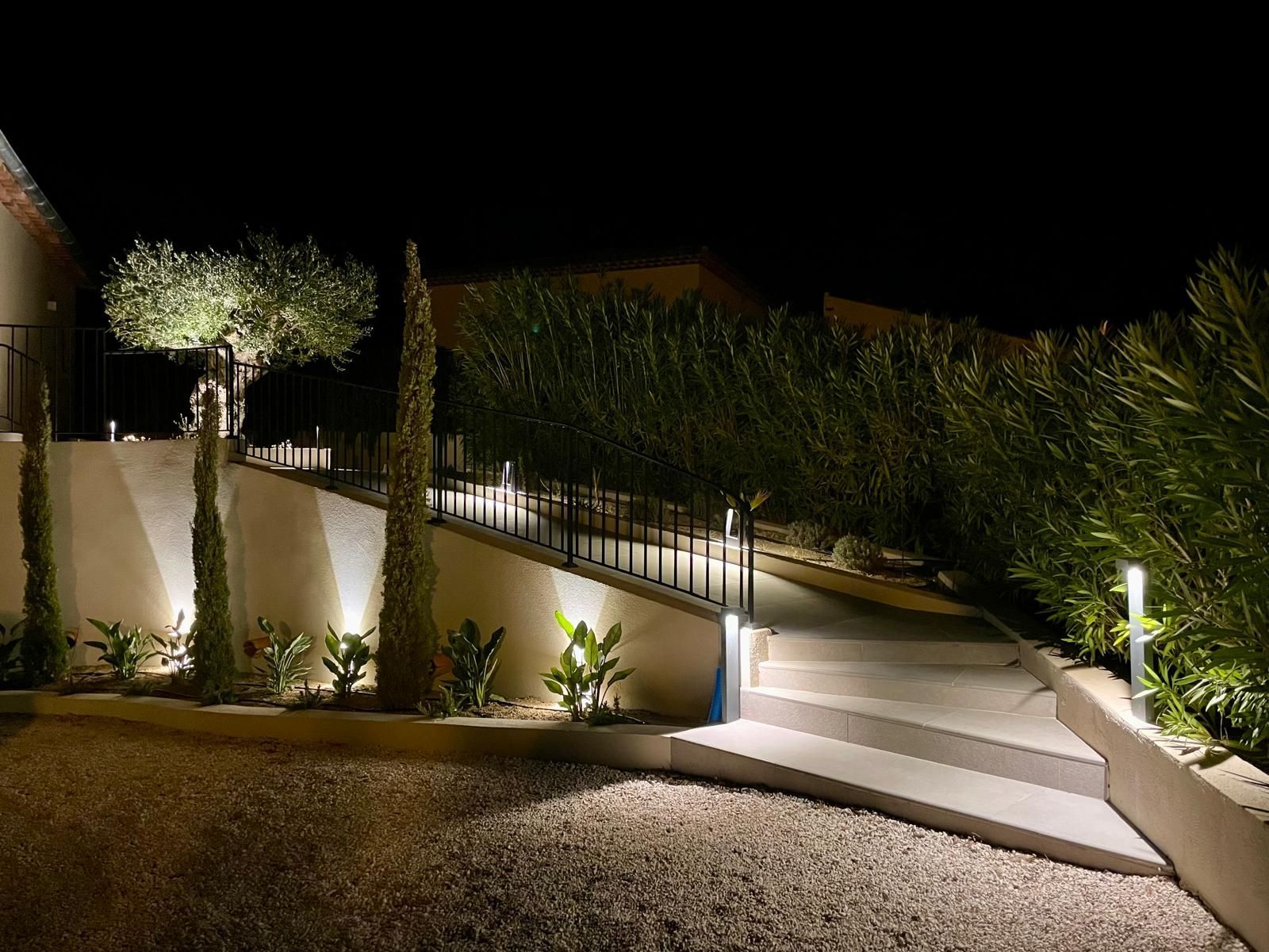 Escalier extérieur nocturne avec murs blancs, rampes noires et aménagement paysager éclairé.