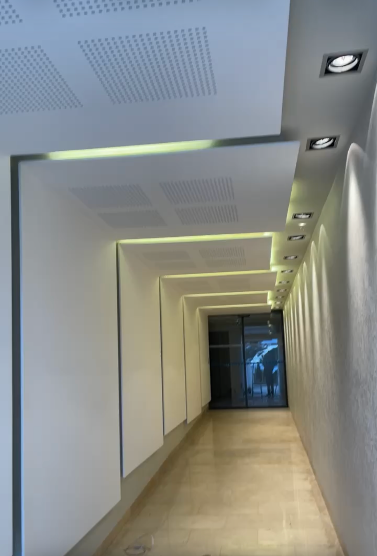 Couloir moderne avec murs blancs, éclairage encastré et sol poli menant à une porte vitrée