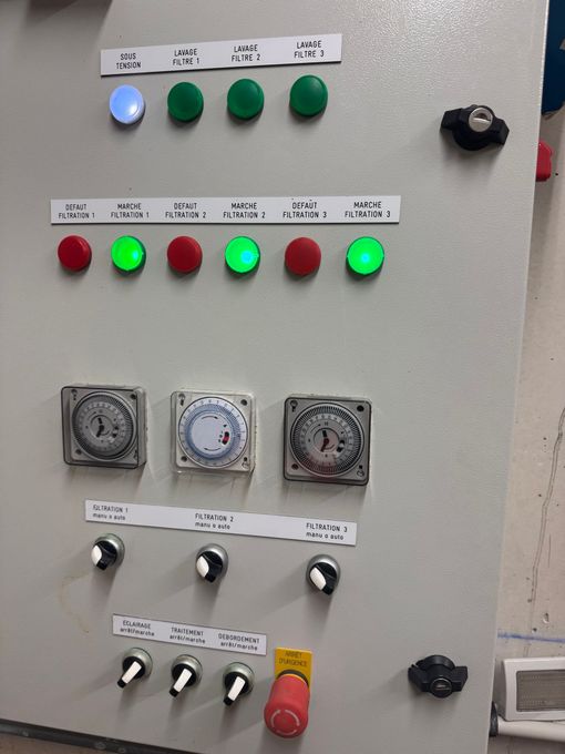 Panneau de commande avec rangées de voyants lumineux colorés, minuteries, interrupteurs et un bouton d'arrêt d'urgence rouge.