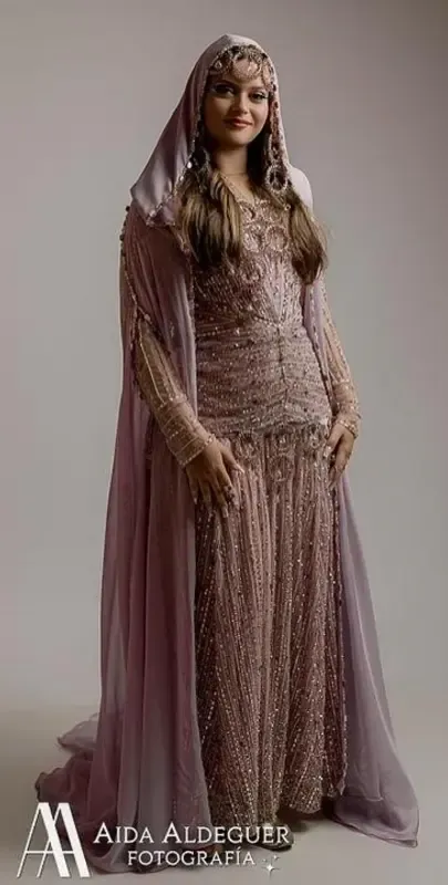 Mujer con un vestido rosa brillante y un pañuelo en la cabeza, de pie frente a un fondo blanco.