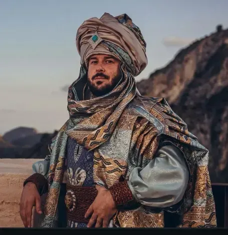 Hombre con elaboradas vestimentas de Medio Oriente, turbante y túnica, frente a un fondo montañoso.