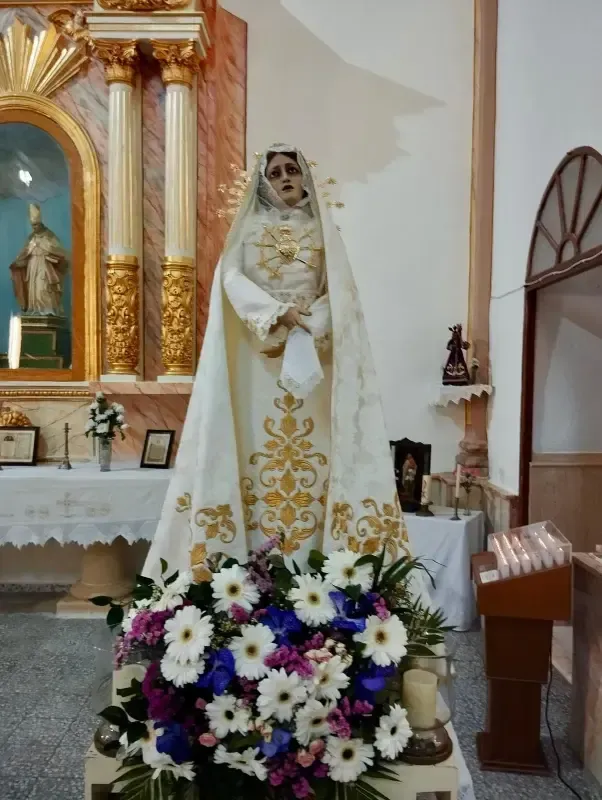 Estatua de una mujer vestida de blanco, de pie en una iglesia, rodeada de flores.