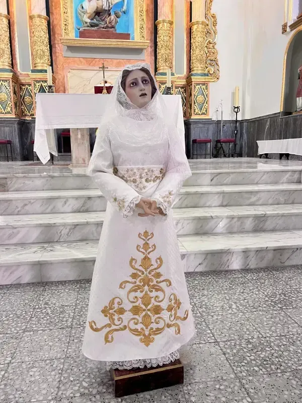 Estatua de una mujer con vestido de novia blanco dentro de una iglesia. El vestido tiene bordados dorados.
