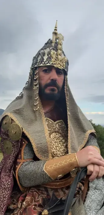 Hombre con elaborada armadura histórica y casco, mirando a la cámara. Al aire libre, cielo nublado.