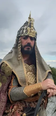 Hombre con elaborada armadura histórica y casco, mirando a la cámara. Al aire libre, cielo nublado.
