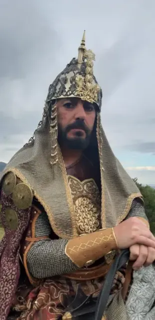 Hombre con elaborada armadura histórica y casco, mirando a la cámara. Al aire libre, cielo nublado.