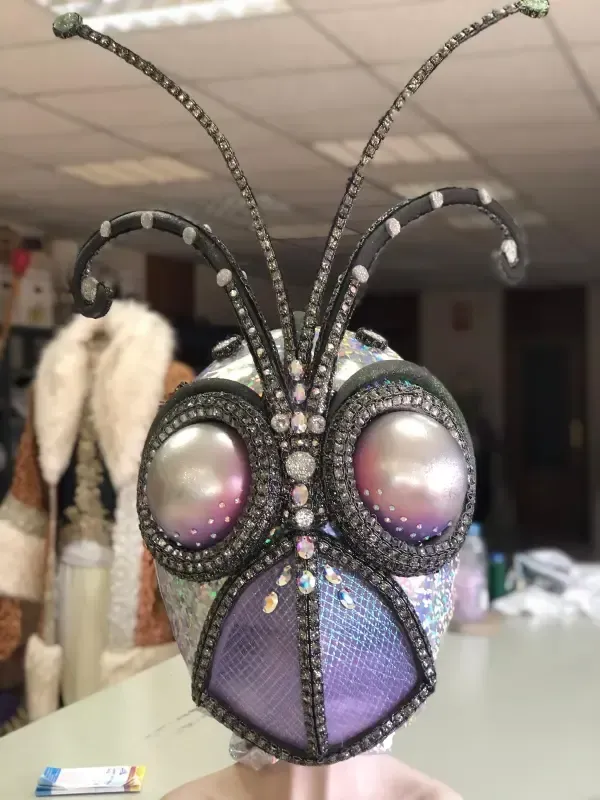 Disfraz elaborado con cabeza de insecto adornada con joyas, con grandes ojos iridiscentes y antenas.