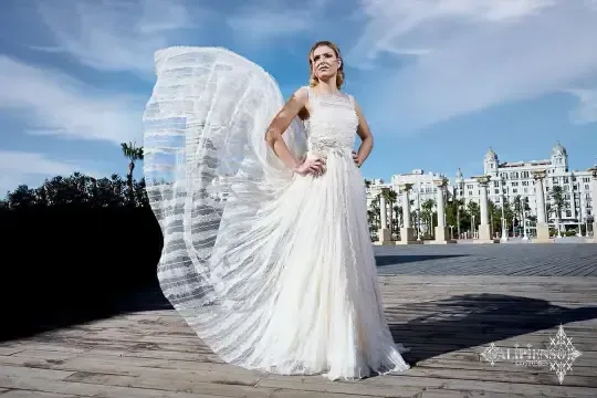 Mujer con vestido de novia y velo suelto, de pie al aire libre con un edificio y un cielo azul de fondo.