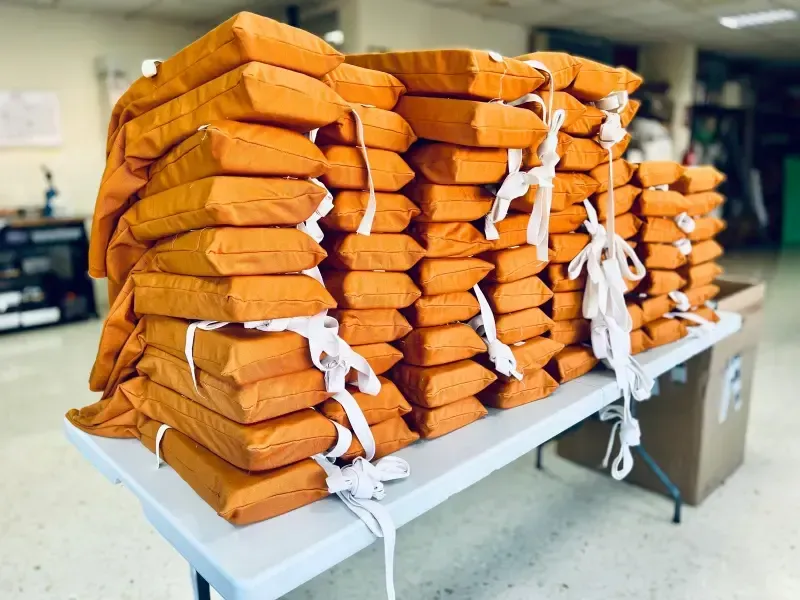 Pilas de cojines naranjas atados sobre una mesa blanca. Interior, probablemente un taller o almacén.