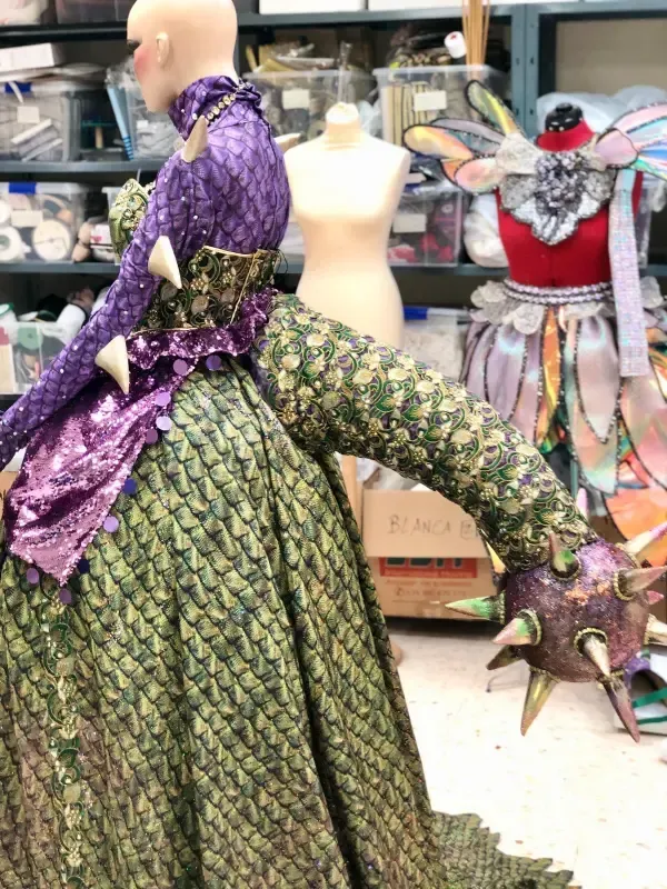 Maniquí con disfraz de dragón: verde y morado, con detalles puntiagudos. Otro maniquí, con temática de hadas, al fondo.