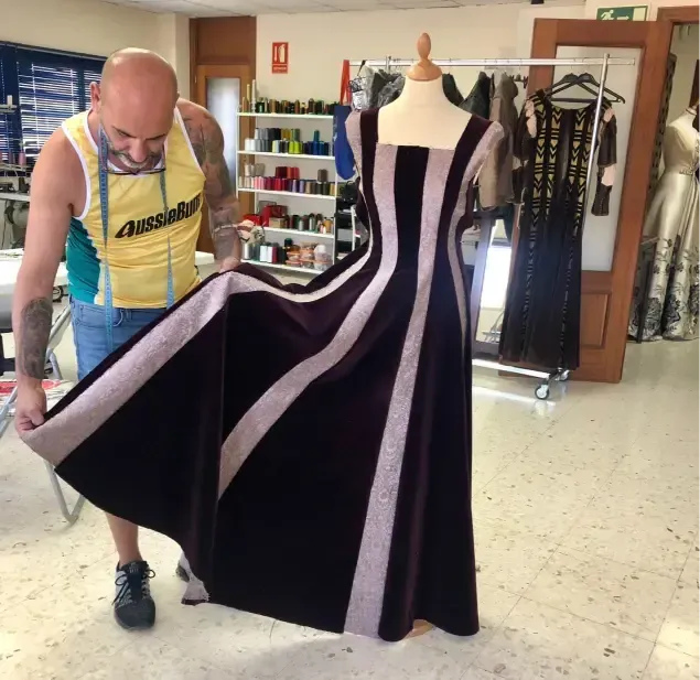 Una persona con una camiseta sin mangas sostiene un vestido morado oscuro con rayas plateadas sobre un maniquí en un taller.