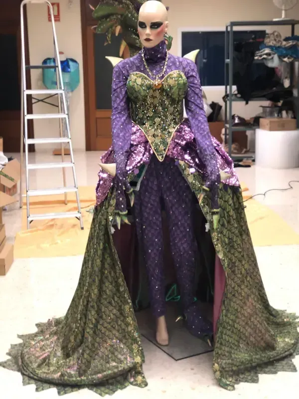 Maniquí con elaborado traje morado y verde con larga cola, en entorno de taller.