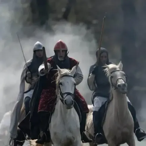 Tres caballeros a caballo avanzan. Uno viste de rojo, los demás llevan cascos de acero. Escena de batalla.