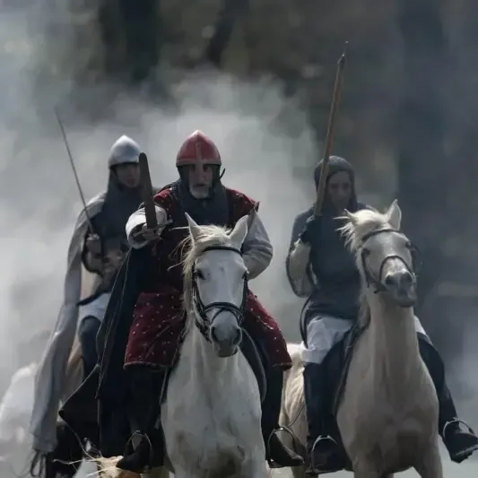 Tres caballeros a caballo avanzan. Uno viste de rojo, los demás llevan cascos de acero. Escena de batalla.