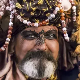 Hombre con turbante adornado y maquillaje facial, sonriendo.