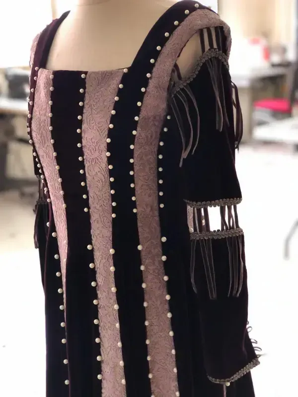 Vestido de terciopelo morado oscuro con paneles rosa claro, adornado con perlas. Mangas con aberturas decorativas.