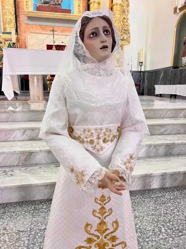Estatua de una mujer con vestido blanco y velo, con las manos entrelazadas. Bordado dorado en el vestido
