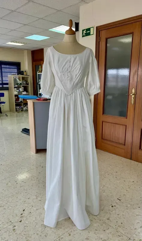 Vestido de algodón blanco sobre un maniquí en una habitación con puerta de madera y muebles de oficina.