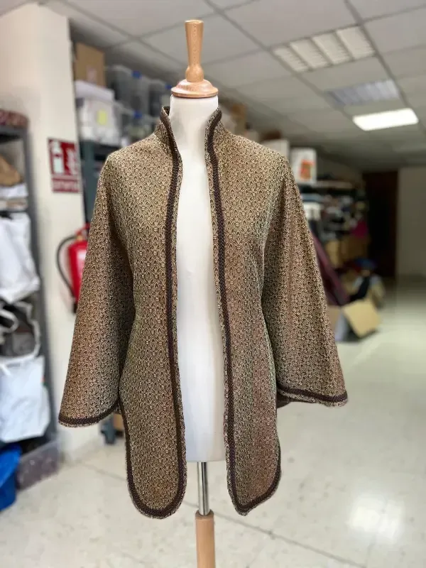 Chaqueta abierta de color canela con ribete marrón sobre un maniquí en una habitación con estantes.