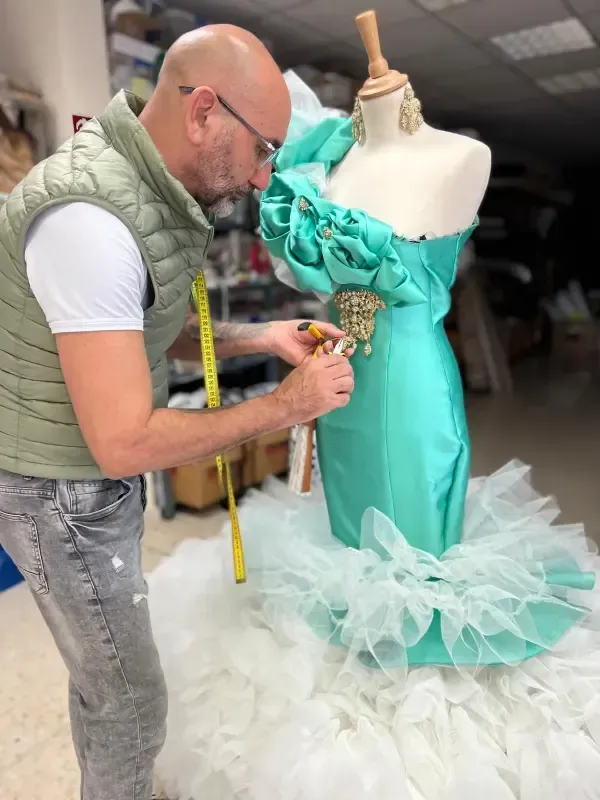 Hombre ajustando un vestido turquesa sobre un maniquí, con falda de tul blanco, dentro de un taller.