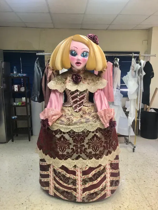 Disfraz de muñeca con peluca rubia y vestido adornado en color rosa, marrón y dorado, de pie en un área detrás del escenario.