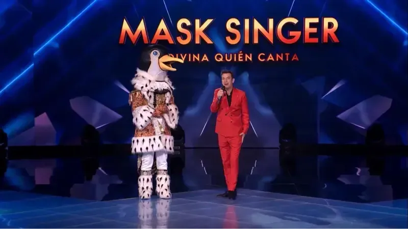 Una persona con un traje rojo se encuentra junto a un pingüino disfrazado en un escenario con un cartel de