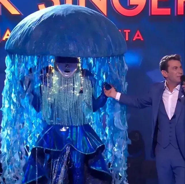 Una persona disfrazada de medusa azul en el escenario con un hombre de traje.