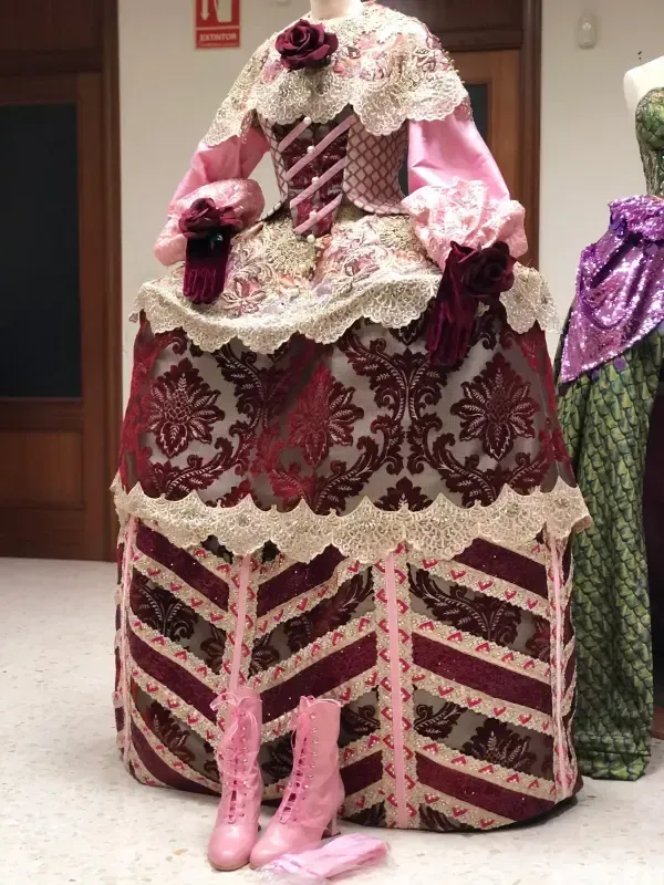Un elaborado traje rosa y burdeos con ribete de encaje, exhibido en un maniquí, con botas rosas en el frente.